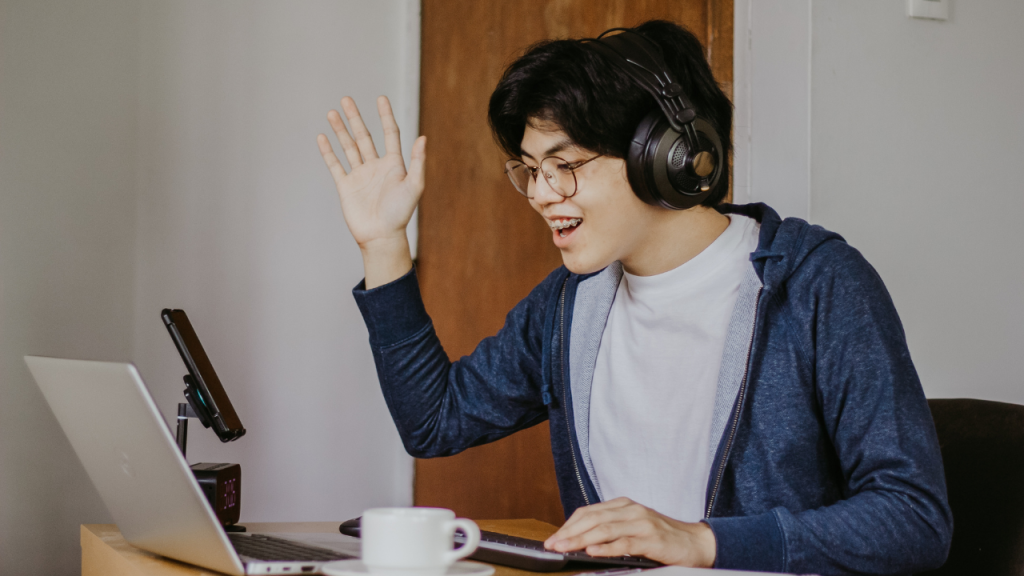A male student raising his hand, facing a laptop to speak in an online class.